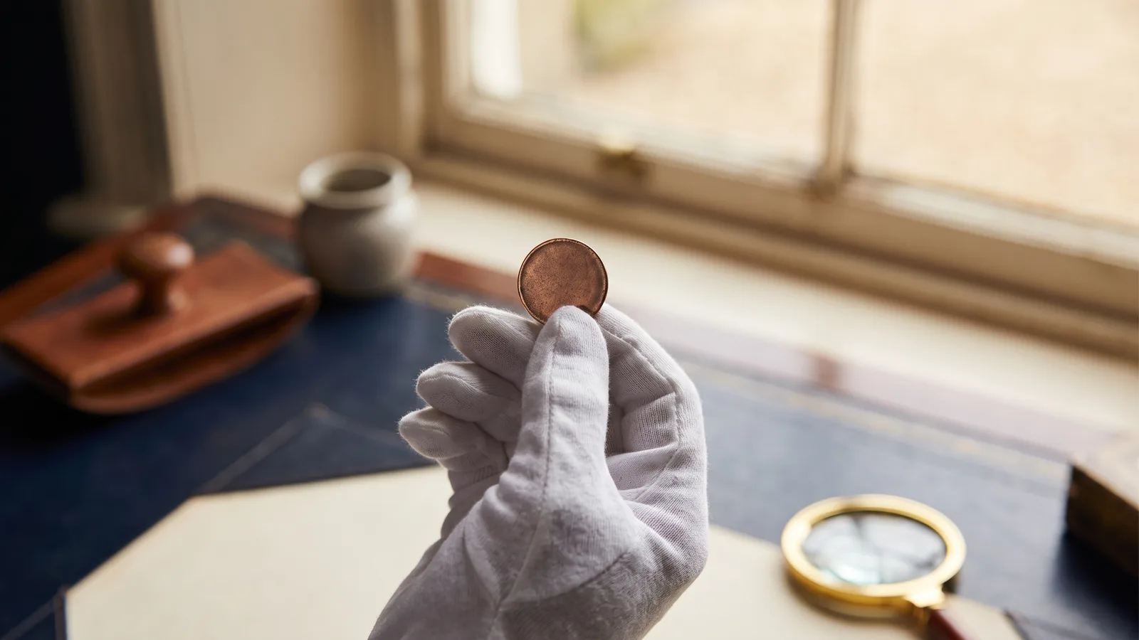 A collector's gloved hand holding a small aged copper token up to warm light