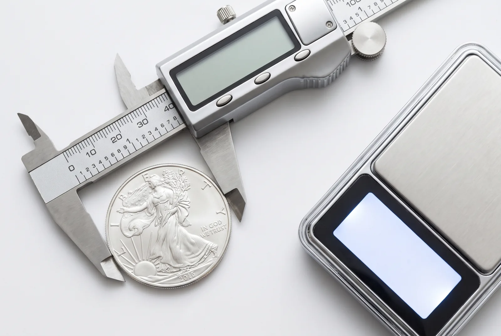 A silver coin on a digital jeweler's scale beside calipers measuring its diameter