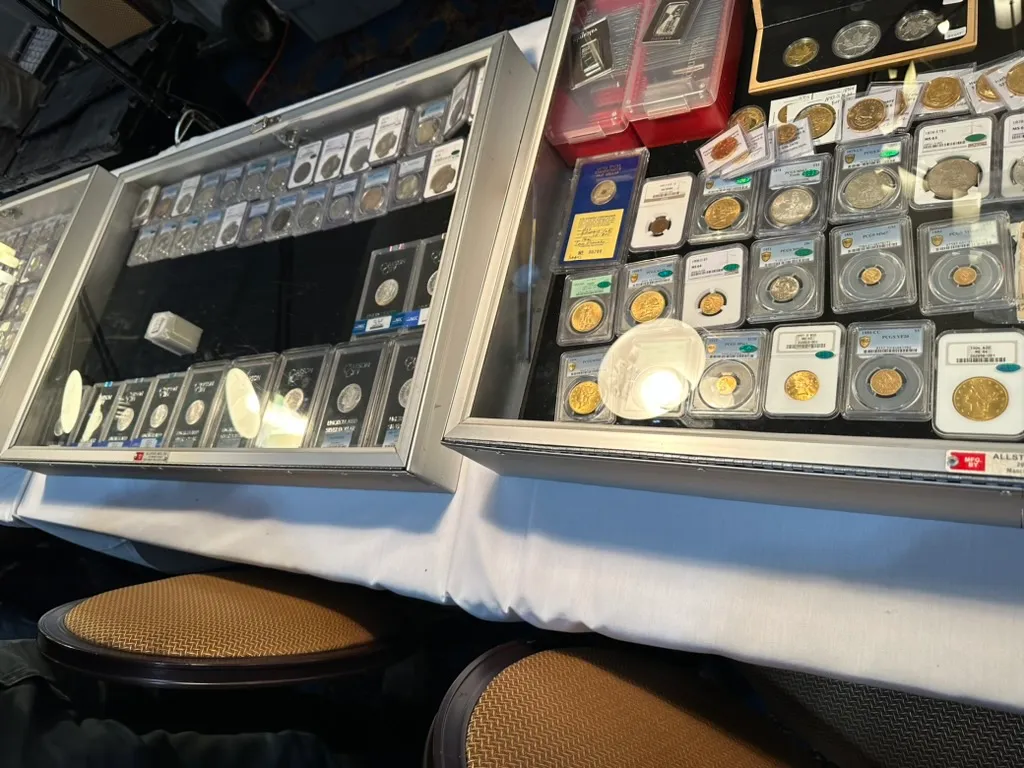 Two glass dealer cases at a coin show holding graded gold and silver coins in labelled plastic slabs, with silver bars behind them