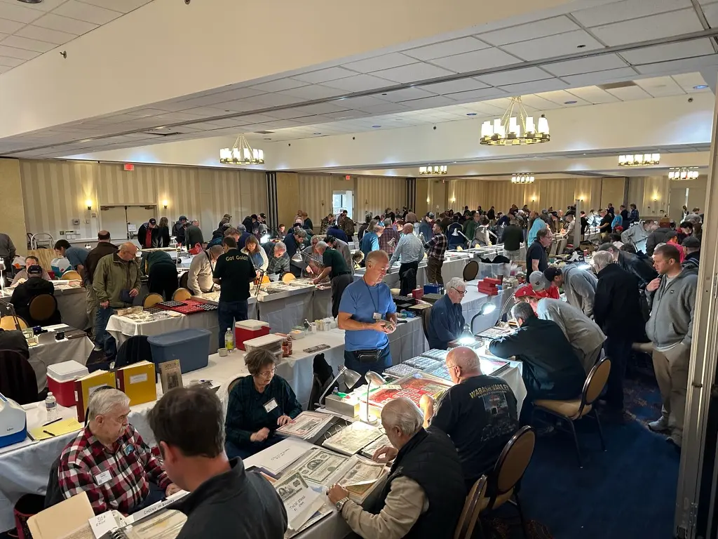 A crowded coin show bourse in a hotel ballroom, with rows of white-clothed dealer tables, desk lamps, and collectors leaning over albums of banknotes